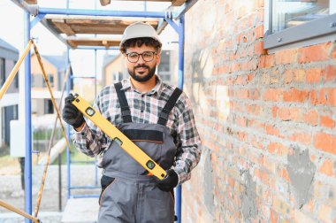 İnşaat işçisi, iş tulumu ve başlık takıyor. İnşaat alanındaki bir iskeleye ruhsal düzeyde bir alet taşıyor.
