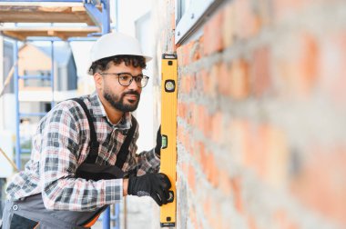 Yeni inşa edilmiş tuğla duvarın ruhsal seviyesiyle dikey hizasını kontrol eden sert şapka ve güvenlik gözlüğü takan profesyonel inşaat işçisi