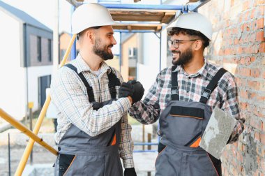 İki farklı miğfer takan, el sıkışan başarılı takım çalışmasını ve şantiyedeki işbirliğini simgeleyen iki farklı müteahhit.