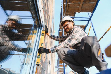 Başlık takan ve eldiven takan yetenekli işçiler inşaat halindeki binanın yeni takılmış penceresini kontrol ediyorlar