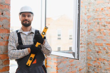 Sert şapkalı ve üniformalı bir adam ruh seviyesinde duruyor. İnşaat alanına tuğladan duvarları olan yeni bir pencere takıyor.