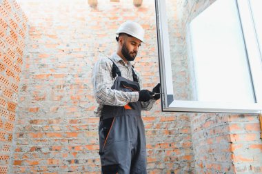 Çalışan adam inşaat başlığı takıyor ve tadilat sırasında tuğla bir duvara yeni bir pencere çerçevesi yerleştiriyor.