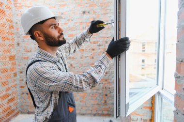Sert şapka ve eldiven takan usta inşaat işçisi, yenileme sırasında tuğla bir duvara yeni bir pencere çerçevesi yerleştiriyor.