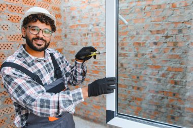 Sert bir şapka, gözlük ve eldiven takan gülümseyen işçi bitmemiş tuğla bir duvara yeni bir pencere çerçevesi yerleştiriyor.