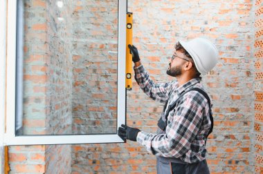 Sert bir şapka ve koruyucu eldiven giyen inşaat işçisi yenileme sırasında yeni pencere çerçevesinin seviyesini kontrol ediyor