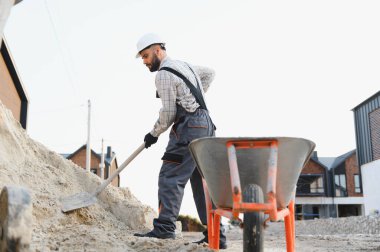 Sert şapkalı ve işçi işçi amelelik yapıyor. İnşaat alanındaki bir el arabasının yanına kürekle kum taşıyor.