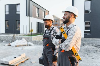 İki yetenekli inşaat işçisi, kasketli ve tulumlu, ellerinde ev inşaatına hazır bir alet var.
