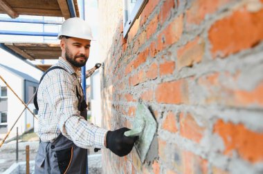Sert şapkalı ve iş kıyafetli genç adam iskelede duruyor, tuğla duvara malayla havan topu uyguluyor.