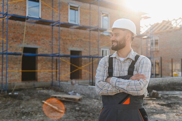 Smiling man in hard hat and overalls standing with arms crossed, proudly looking at new house construction