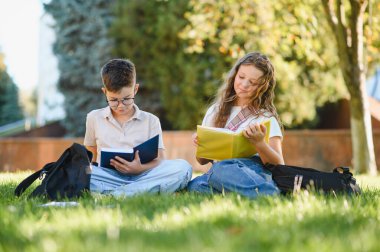 Yeşil çimenlerde oturan genç öğrenciler, okul çantalarıyla birlikte kitap okumaya odaklanmış, öğrenim ve eğitimi teşvik ediyorlar.