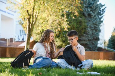 İki genç öğrenci yeşil çimlerde oturuyor, güneşli bir günde üniversite binasında sırt çantalarındaki okul malzemelerini paylaşıyorlar.