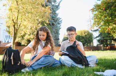 Öğrenciler eğitime hazırlanıyor, okul çantalarını ve kitapları topluyor ve dışarıda yeşil çimlerin üzerinde oturuyorlar.