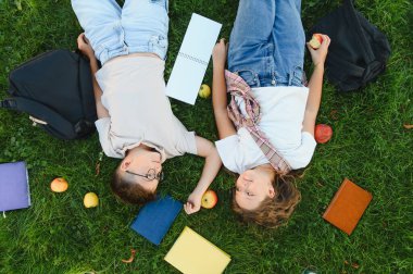 Yeşil çimenlerde uzanan iki genç öğrenci, el ele tutuşurken kitaplar ve sırt çantasıyla mola veriyorlar.