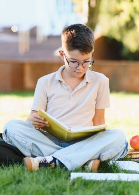 Gözlüklü bir çocuk çimenlerde oturmuş kitap okuyor, dışarıda ödev yapıyor ve kitap okuyor.