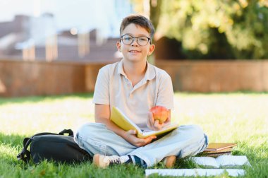 Yeşil çimenlerde oturan, kitap okuyan ve parkta elma yiyen gülümseyen çocuk. Öğrenme kavramı