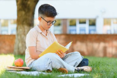Gözlüklü genç bir erkek öğrenci okul binasının yakınındaki yeşil çimlerde ders kitabı okuyor, tatilde öğreniyor.