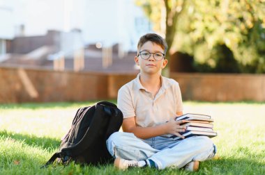 Yeşil çimlerin üzerinde oturan genç bir öğrenci, elinde bir yığın okul kitabı ve yakında öğrenmeye hazır bir sırt çantası var.