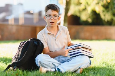 Gözlüklü genç bir çocuk yeşil çimenlerde oturuyor, elinde kitaplar, yanında sırt çantası, şaşkınlığını dile getiriyor.