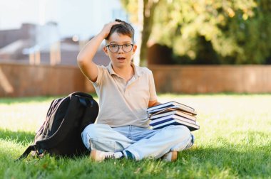 Öğrenci gözlüklü çocuk çim üzerinde kitap ve sırt çantasıyla oturuyor. Okul ödevleri konusunda kafası karışmış ve stresli görünüyor.