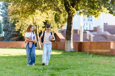 İki okul çocuğu yeşil bir kampüs bahçesinde yürüyorlar, kitap taşıyorlar, okula gidiyorlar, öğrenmeyi ve arkadaşlığı simgeliyorlar.