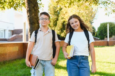 Kitapları ve sırt çantaları olan mutlu çocuklar, güneşli bir günde kampüste dururken gülümsüyorlar.