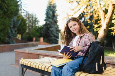 Genç liseli kız modern bir bankta oturmuş kitap okuyor. Açık havada öğrenmek, okul parkında eğitimin tadını çıkarmak.