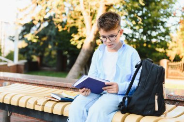 Öğrenci parktaki bankta oturmuş kitap okuyor, ödevlere hazırlanıyor ve ders çalışıyor. Sırt çantası yanında.