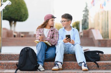 Çocuklar okul merdivenlerinde oturuyor, ellerinde cep telefonları ve sohbet ediyorlar, arkadaşlıklarını ve modern öğrenci yaşamlarını gösteriyorlar.