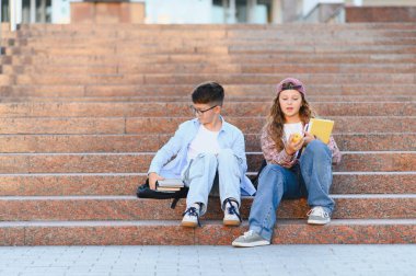 Öğrenciler granit merdivenlerde oturup kitaplara ve dijital tablete bakıyorlar. Sıradan kıyafetler giyiyorlar.