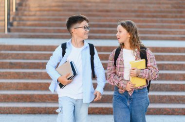 Sırt çantaları ve kitaplarıyla birlikte yürüyen, gülümseyen ve okuldan ayrılırken konuşan iki genç öğrenci.