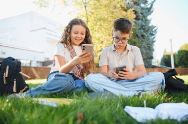Okul çocukları kampüs çimlerinde oturuyor, cep telefonlarıyla uğraşıyorlar, derslere kısa bir ara veriyorlar.
