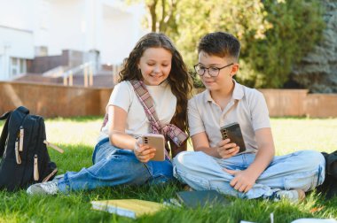 Mutlu okul çocukları çimenlerde oturuyor, telefonlara bakıyor, dersten sonra birlikte vakit geçiriyorlar, modern teknolojiyi bağlantı için kullanıyorlar.