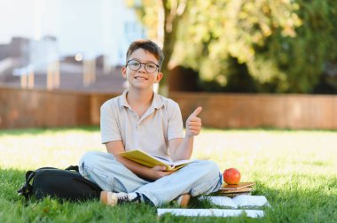 Çimlerin üzerinde oturan mutlu genç çocuk kitap, sırt çantası ve elma okuyor. Eğitim, öğrenme ve başarı kavramı