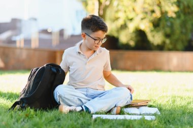Sırt çantalı ve kitaplı genç bir erkek öğrenci yeşil çimlerin üzerinde oturuyor, okul binasının dışında okuyor ve öğreniyor.
