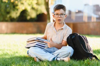 Öğrenci elinde bir yığın kitap ve sırt çantasıyla kampüsteki yeşil çimlerin üzerinde oturarak çalışmaya hazırlanıyor.