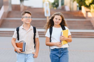 İki okul çocuğu, bir oğlan ve bir kız, okul kampüsünde geziyorlar, kitap taşıyorlar ve sırt çantaları takıyorlar.