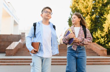 İki genç öğrenci, bir erkek ve bir kız, okul binasının önünde birlikte yürüyorlar, sırt çantaları ve kitaplar taşıyorlar.
