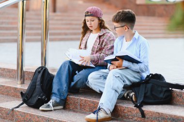 Genç öğrenciler ödevleri tartışıyor ve kampüs basamaklarında kitap okuyor, eğitimi, arkadaşlığı ve çocukluğu temsil ediyor.