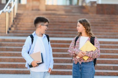 İki genç öğrenci, bir erkek ve bir kız, açık bir merdivende duruyorlar, kitap ve sırt çantaları taşırken konuşup gülüyorlar.