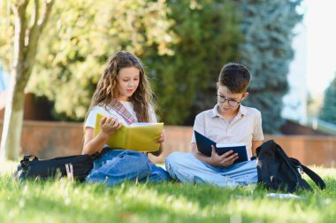 Genç öğrenciler birlikte öğreniyor ve eğitimin tadını çıkarıyorlar, okul tatilinde sırt çantalarıyla yeşil çimlerin üzerinde oturuyorlar.