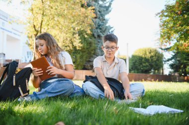 Yeşil çimenlerde oturan öğrenciler kitap okuyor ve derslere hazırlanıyor, açık havada eğitimin tadını çıkarıyorlar.