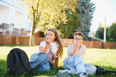 İki okul çocuğu yeşil çimlerde oturuyor, taze elmaların tadını çıkarıyor ve okul binasının dışında vakit geçiriyorlar. Sırt çantaları ile dinleniyorlar.