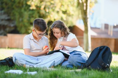 Okul çocukları yeşil çimlerde oturuyor, okuyor ve öğreniyor, çocuk elma tutuyor, kız kitabı işaret ediyor.