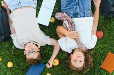 İki neşeli ilkokul çocuğu, bir oğlan ve bir kız, yeşil çimlerde, elma ve okul malzemeleriyle mutlu bir şekilde dinleniyorlar.