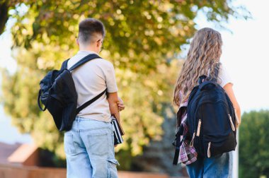 İki öğrenci, bir oğlan ve bir kız, sırt çantaları ve kitaplarıyla, güneşli bir günde dışarı çıkıp, okula gidiyorlar.
