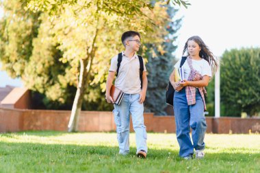 Okul çocukları yeşil bir çimenlikte yürüyorlar, çantalarını ve kitaplarını doğal ışık altında taşıyorlar, sınıfa gidiyorlar.