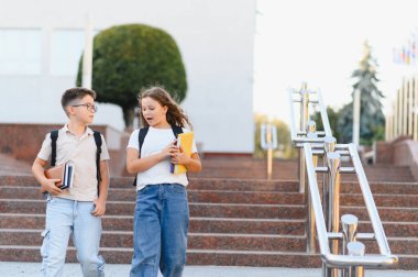 Okul çocukları sırt çantaları ve ders kitaplarıyla merdivenlerden iniyor, okul binasının önünde sohbet ediyorlar.