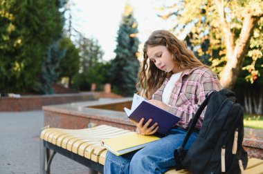 Parktaki bankta oturan genç bir kız. Sırt çantalı bir kitaba odaklanıyor. Öğrenim ve eğitim kavramı