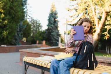 Sırt çantası ve kitaplarla bankta oturan liseli kız, eğitim kampüsünde ders çalışmaya hazırlanıyor.