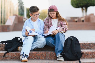 İki genç öğrenci, erkek ve kız, okul merdivenlerinde oturuyor, okul ödevlerini öğreniyor ve tartışıyorlar, sırt çantaları yakınlarda.